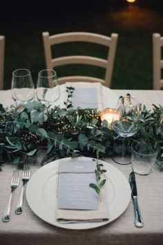 the table is set with white plates and silverware, greenery and candlelight