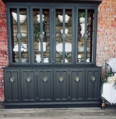 an old china cabinet with glass doors and flowers on the top, in front of a brick wall