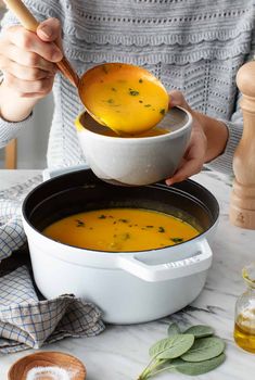 a woman is holding a spoon over a bowl of soup on a table with other ingredients