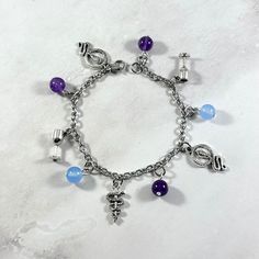 This Hygeia themed, charm bracelet is the perfect accessory to showcase your love for the Greek gods in a subtle way.  Hygeia is the Greek goddess of good health and is usually depicted with a snake and a cup.  Charms included: Snake Cup Rod of Asclepius Snake Cup 3 amethyst and 2 blue quartz beads are also included between each charm.  Comes in three sizes: Small: 6.5 inches (with extra rings at the end to expand up to 7.5 inches.) Standard: 7.5 inches Large: 8.5 inches Secures with a lobster clasp. Bracelet and charms are lead and nickel free. Comes in a jewelry box so it would be ready to be given as a gift! Message me of you would like to substitute any of the charms. If you have any questions or need more quantities than are currently available, please message me. Looking for a custom Hygiea Greek, Greek Goddess Bracelet, Rod Of Asclepius, The Greek Gods, Greek Mythology Jewelry, Cup Charms, Mythology Jewelry, Goddess Bracelet, Clasp Bracelet