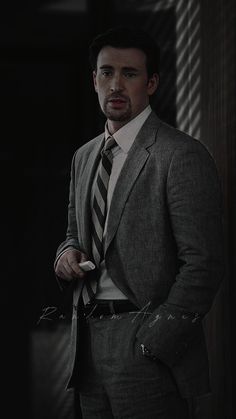 a man wearing a suit and tie standing in front of a dark wall with his hands in his pockets