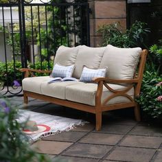 a wooden couch sitting on top of a patio next to some bushes and flowers in front of a black gate