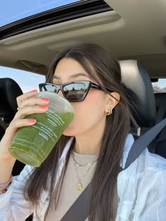 a woman drinking from a green drink in the back seat of a car while wearing sunglasses