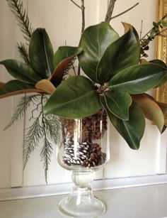 a vase filled with green leaves and pine cones on top of a white table next to a mirror