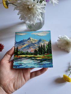 a hand holding a small painting in front of flowers