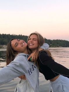 two young women standing next to each other in front of the water with trees behind them