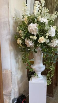 a white vase filled with lots of flowers on top of a table next to a wall