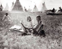 an old black and white photo of two children sitting on the grass in front of teepees