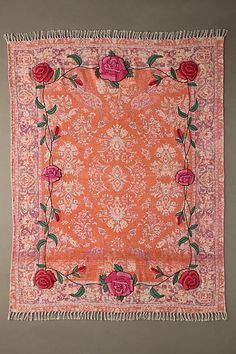 an orange and pink square rug with roses on the border, hanging from a gray wall