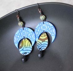 These celestial earrings feature beautiful blue and silver crescent polymer clay components. The crescents have a striped floral design. Dangling below are teardrop shaped enamel components with a geometric design in blue and green. They are accented with Czech glass beads, copper spacers and stone beads. Dangling below are brass ball charms. These unique earrings measure approx. 3 5/8 inches long dangling on niobium earring hooks. All pieces come boxed ready for gift giving or keeping for yours Blue Celestial Pierced Earrings, Celestial Blue Pierced Earrings, Blue Celestial Nickel-free Earrings, Blue Crescent Moon Charm Earrings, Blue Crescent Earrings As A Gift, Blue Round Earrings With Moon Charm, Blue Crescent Jewelry With Sun And Moon Design, Unique Sun And Moon Design Drop Earrings, Blue Dangle Earrings With Moon Charm