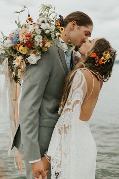 a bride and groom kissing in front of the ocean with flowers on their head, holding each other's hands