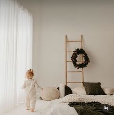 a toddler standing on the edge of a bed in front of a window with white curtains