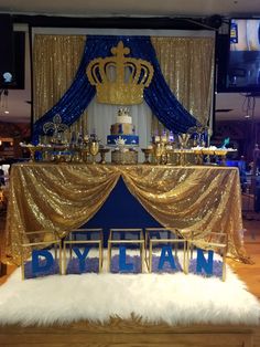 a gold and blue dessert table with a crown on top, surrounded by white fluffy rugs