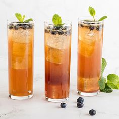 three tall glasses filled with iced tea and blueberries on a marble table next to mint sprigs