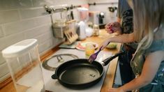 two people are cooking in a kitchen on the stove top and one is using a frying pan