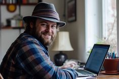 a man sitting at a desk with a laptop computer in front of him and wearing a hat