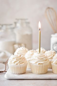 cupcakes with white frosting and a single candle on a tray in front of jars