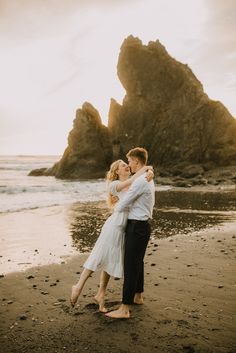 a man and woman hugging on the beach with rocks in the background at sunset time