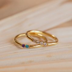 two gold rings with different colored stones on top of a wooden table next to each other