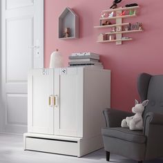 a pink room with a gray chair and white dresser next to a shelf that has books on it