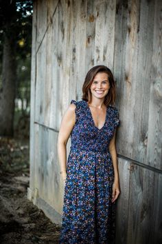 a woman standing in front of a wooden wall wearing a blue dress and smiling at the camera