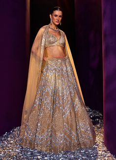 Embrace elegance with the Gold Sequins Embroidered Bridal Lehenga Set, a true masterpiece of craftsmanship and design. Crafted from soft net fabric, the golden lehenga boasts an intricately embroidered pattern, featuring a stunning blend of silver and gold sequins, crystals, and bugle beads that create a captivating sparkle from every angle. Teamed with a blouse equally adorned with detailed embroidery, this ensemble radiates luxury and sophistication. The look is completed with a sheer dupatta that adds an ethereal touch of elegance. This perfect blend of traditional artistry and modern style makes the ensemble ideal for making a memorable entrance. Perfect for a bride or bride-to-be, this lehenga set ensures you shine with grace and beauty on your special day. Composition : Lehenga, Blou Gold Floor-length Sharara With Resham Embroidery, Traditional Gold Embroidered Gown For Festive Season, Floor-length Sharara With Gold Embroidery For Diwali, Elegant Designer Lehenga With Gold Embroidery, Gold Lehenga With Mirror Work For Eid, Gold Sharara With Mirror Work For Wedding, Festive Gold Lehenga With Mirror Work, Gold Mirror Work Sharara For Wedding, Gold Sharara With Gold Embroidery For Festivals