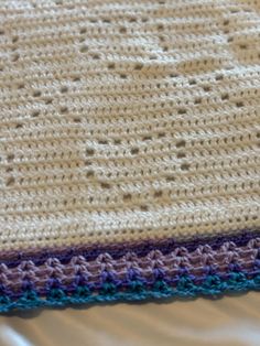 a crocheted blanket laying on top of a white bed cover with blue and purple trim