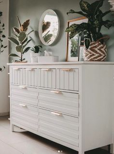 a white dresser sitting next to a mirror and potted plants