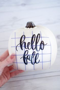 a hand holding up a white pumpkin with the words hello fall painted on it in black ink