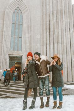 three young women standing in front of a church on a snowy day with one taking a photo