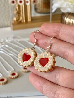 a person is holding some kind of heart shaped cookie earrings in their hand and there are other cookies on the table behind them