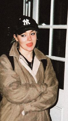 a woman wearing a hat and jacket standing in front of a window with her arms crossed