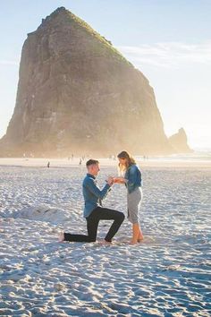 a man kneeling down next to a woman on the beach with a mountain in the background