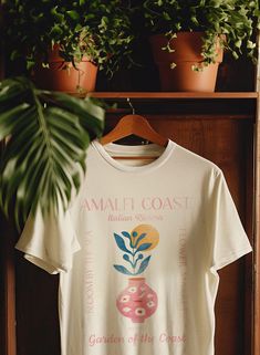 a white t - shirt hanging on a wooden shelf next to potted plants and a plant