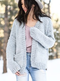 a woman standing in the snow wearing a gray hat and cardigan sweater with pockets