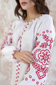 a woman wearing a white shirt with red designs on the chest and sleeves, standing in front of a wall