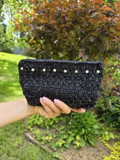 a hand holding up a black purse with pearls on the front and bottom, in front of some bushes