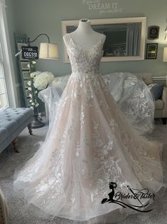 a wedding dress on display in a room with furniture and flowers behind it, the bride is wearing a white lace appliqued gown