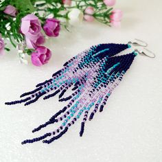 purple and blue beaded fringe earrings on a white surface next to pink flower stems