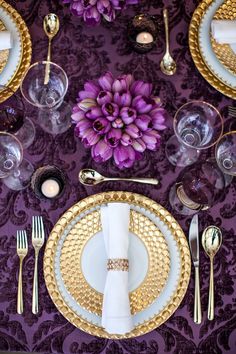 the table is set with purple flowers and gold place settings, silverware, and napkins