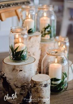 candles are lit in glass jars with greenery on them, along with birch logs