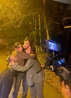 three people hugging each other in front of a camera set up on the side of a road