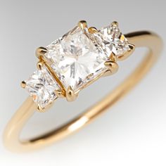 three stone diamond ring in yellow gold with two princess cut diamonds on each side, set against a plain white background