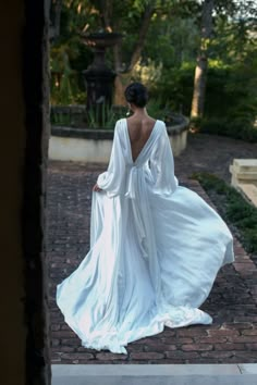 the back of a woman in a white dress walking down a brick walkway with trees and bushes behind her