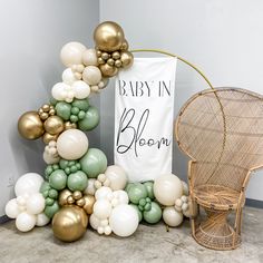 a baby in bloom sign next to a chair and balloon arch with gold, white, and green balloons