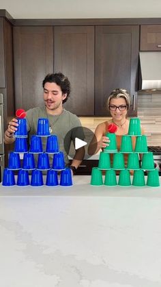 two people standing in front of a kitchen counter with blue and green cups stacked on top of each other