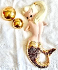 a mermaid figurine sitting on top of a bed next to two golden balls