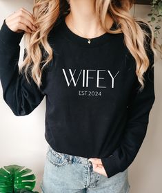 a woman wearing a black sweater with the word wife printed on it, standing in front of a potted plant