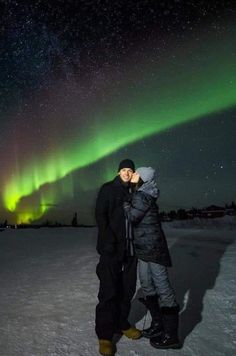 two people are standing in the snow with an aurora light behind them and one person is kissing