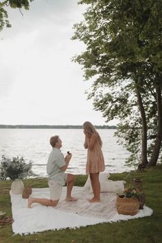 a man and woman sitting on a blanket near the water, talking to each other